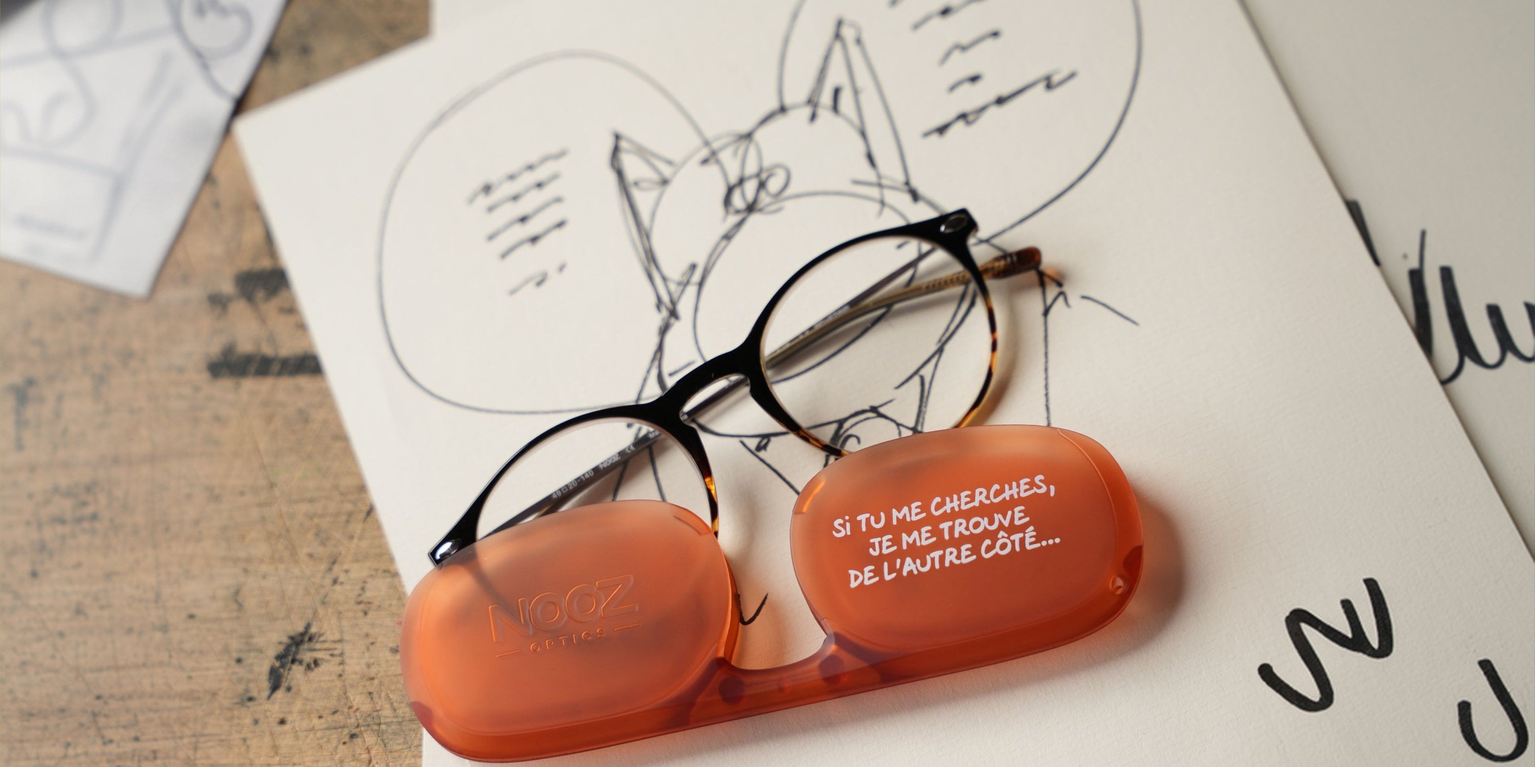 Lunettes NOOZ CRUZ posées sur un croquis du Chat, à côté de l'étui orange